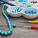 Two bead reamers in use on a work surface beside a blue bead strand and a tray of beads