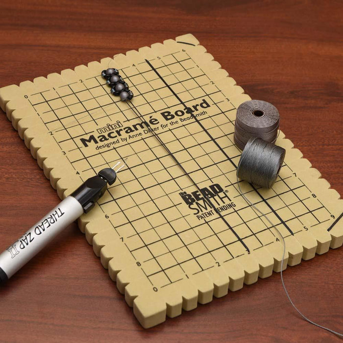 Mini macrame board on a wood table with beads, spools of cord and a thread zip tool beside it