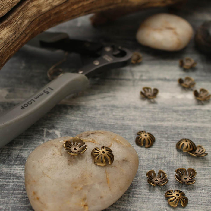 Antiqued brass flower bead caps on smooth stones with loop-making pliers and driftwood in a styled workspace
