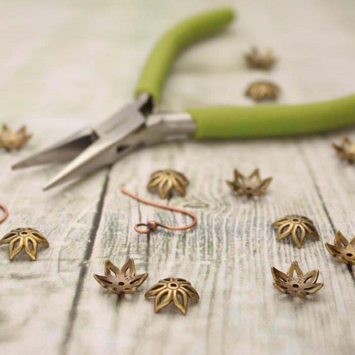 Antiqued brass flower bead caps on weathered wood beside green-handled pliers and a copper ear wire