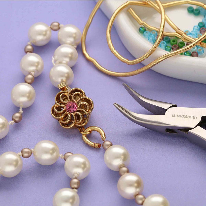 Gold French wire beside pearl necklace with flower clasp, pliers and a dish of seed beads on a purple background
