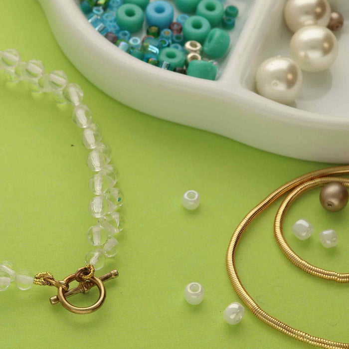 Gold French wire lengths on a green surface next to a clear bead necklace with toggle clasp and a dish of colorful beads