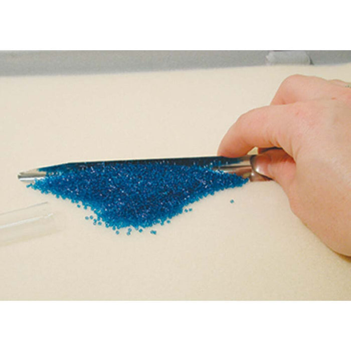 Hand using the Scoop-EEZ bead scoop to gather a pile of tiny blue seed beads from a flat surface