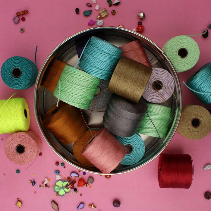 Overhead view of a tin filled with S-Lon nylon cord spools in many colors on a pink surface with loose beads