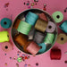Overhead view of a tin filled with S-Lon nylon cord spools in many colors on a pink surface with loose beads