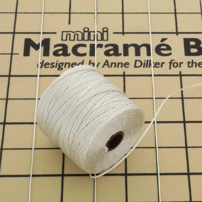 Spool of S-Lon cream nylon bead cord resting on a mini macrame board with cord strands pinned across it