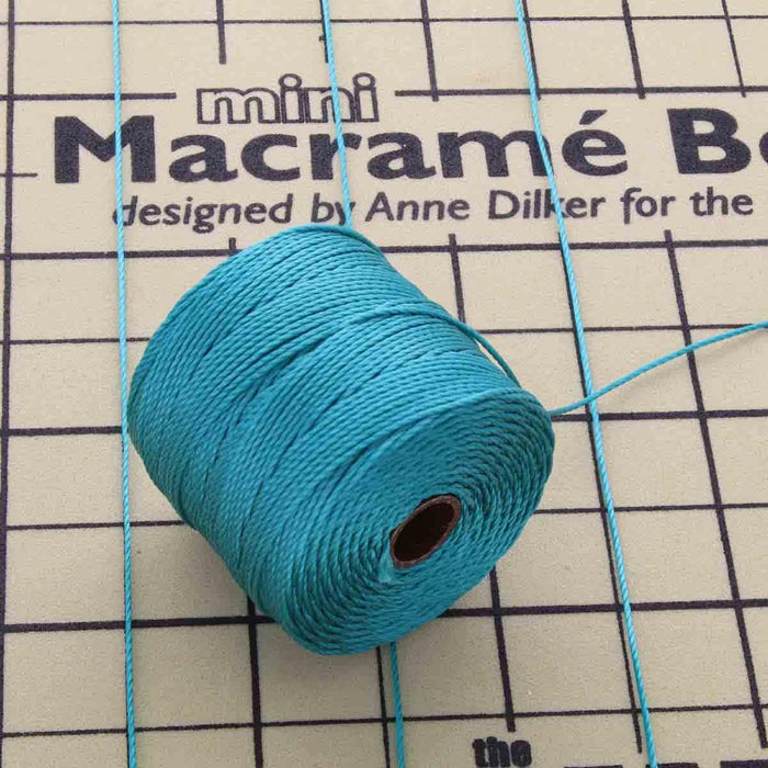 Teal S-Lon nylon bead cord spool resting on a mini macrame board with cord strands pinned across it