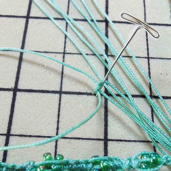 T-pin holding green macramé cords with green seed beads on a gridded work board