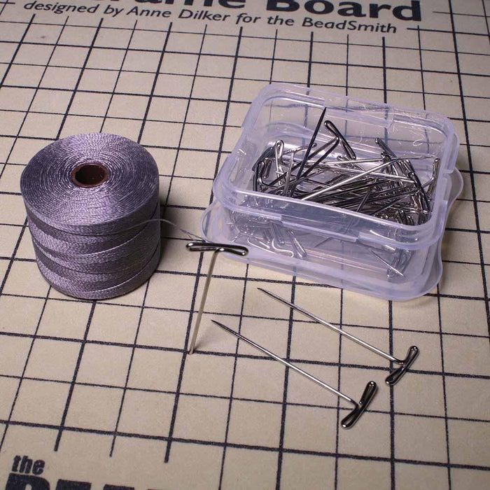 Open container of T-pins with loose pins and a spool of gray cord on a printed macramé board