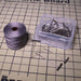 Open container of T-pins with loose pins and a spool of gray cord on a printed macramé board