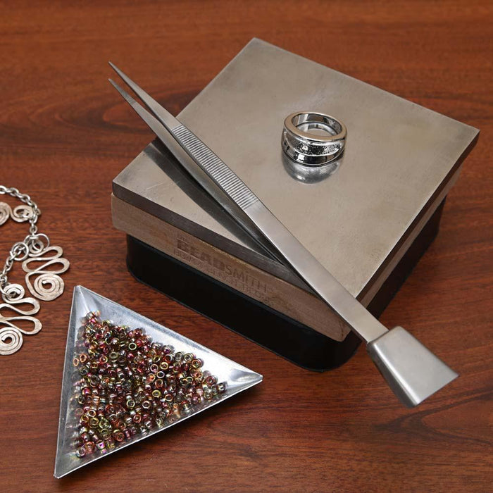 Tweezers with shovel resting on a steel bench block beside a ring, seed bead tray and wire jewelry
