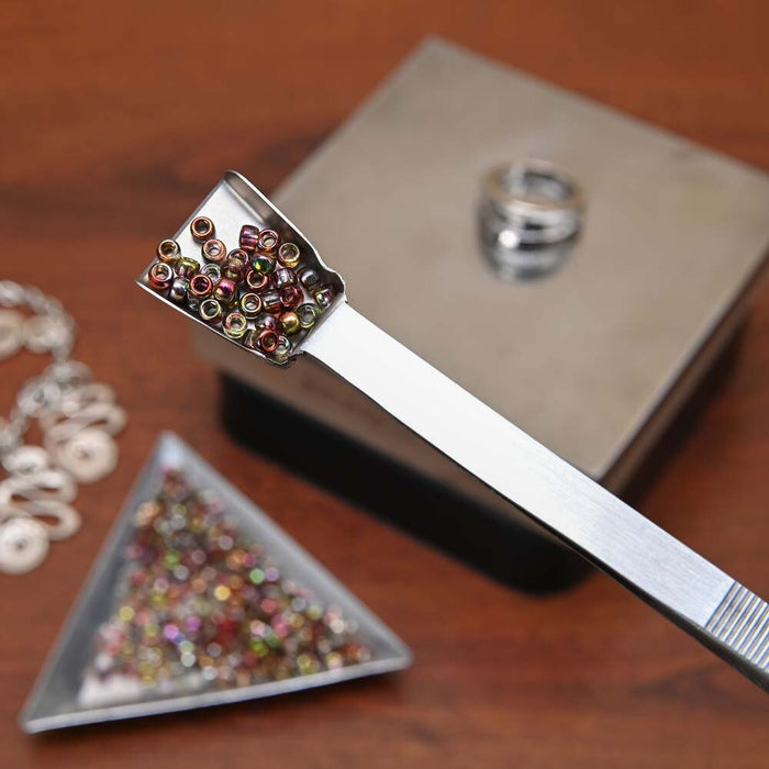Shovel end of the tweezers scooping a small pile of seed beads, with a tray of beads below