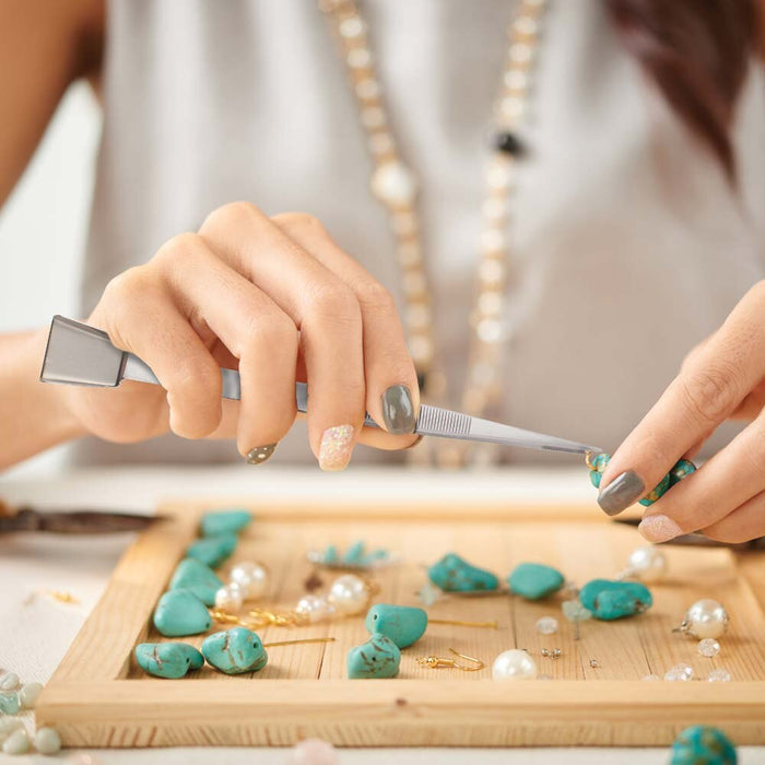 Person using the tweezers to pick up a turquoise-colored stone bead at a bead sorting tray while making jewelry